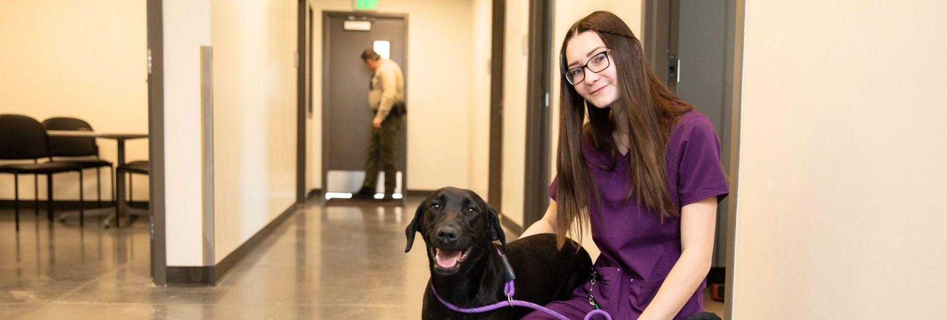 Woman with long brown hair in purple scrubs kneeling down with a black dog
